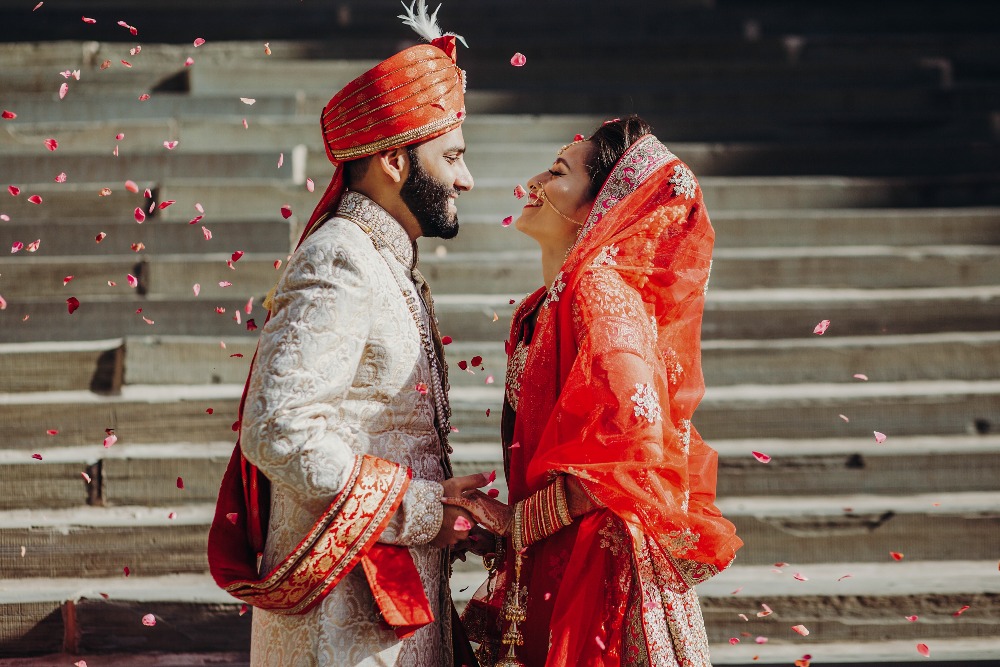 Indian groom dressed in white Sherwani and red hat with stunning bride in red lehenga stand and hold each hands walking outside